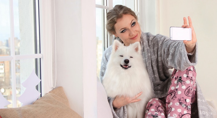 Millenial in pink pajama pants and a grey sweater taking a selfie with a white medium sized furry dog