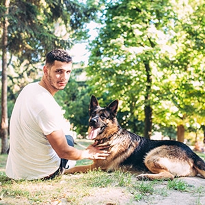 Man outside with German Shepard, both sitting on the ground in the woods