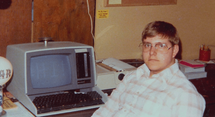 Guy in classes and plaid shirt sitting at computer in the 80's